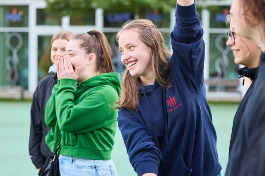 Vier Jugendliche stehen drau&szlig;en vor einem Geb&auml;ude. Ihr Oberk&ouml;rper . Ein M&auml;dchen hebt ihren Arm hoch und lacht.