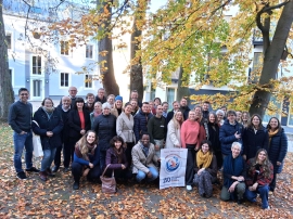 Männer und Frauen stehen für ein Gruppenbild in einem Innenhof vor Bäumen, die ihr Herbstlaub verloren haben.