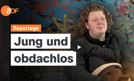 Ein junger Mann mit schwarzem Pullover und Wolldecke sitzt vor einer Wand mit Graffiti und blickt zur Seite.