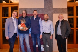 Fünf Männer stehen vor einer grau-weißen Wand. Einer davon hält einen Blumenstrauß in den Händen.