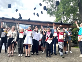 Junge Männer und Frauen stehen vor einem Gebäude. Sie halten ein Batt Papier in der einen Hand und werfen eine Mütze mit der anderen Hand in die Luft.