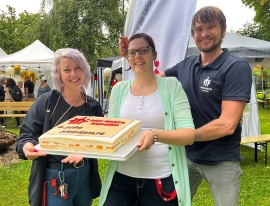 Zwei Frauen und ein Mann stehen auf einer Wiese, im Hintergrund Gartenpavillions und Sitzbänke. Sie halten einen Kuchen mit Aufschrift "10 Jahre Jugendhilfe".