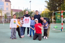 Luke Kelly steht it Kindern und Erwachsenen auf einem Sportplatz. Ein Erwachsener hält ein T-Shirt hoch, auf dem "700 Kilometer für Kinder in Kenia" steht.