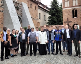Piotr Motwicki in schwarzem Rock und weißem Übergewand steht mit Männer, Frauen und Jugendlichen vor einer Backsteinkirche.