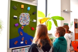 Drei Schüler stehen vor einer grünen Tafel, auf der im Kreis bunte Papierboote aufgeklebt sind. In der Mitte ein grauer Filzkreis mit kleinen bunten Papierkugeln.