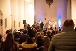 In einer Kirche sind im Vordergrund Menschen in Kirchenbänken  von hinten zu sehen. Vorne steht eine Sängerin und am Altar zwei Priester in weißen Gewändern.