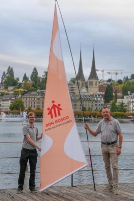 Vor einem Fluss steht ein junger und ein älterer Mann mit einem Segel mit Don Bosco Logo