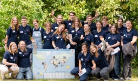23 junge Frauen und Männer mit Don-Bosco-Volunteers-T-Shirt stehen oder hocken für ein Gruppenbild in einem Park. In der Mitte steht eine Weltkarte.