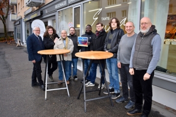 Mehrere Personen sehen vor einem Ladengeschäft und blicken in die Kamera. Vor ihnen stehen zwei Stehtische.
