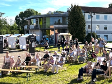 Menschen sitzen bei sonnigem Wetter vor in Liegestühlen vor dem Don Bosco Zentrums-Gebäude.