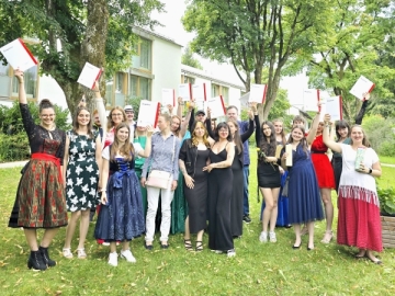Festlich gekleidete junge Menschen stehen auf einer Wiese und halten Dokumente mit in ihren Händen hoch.