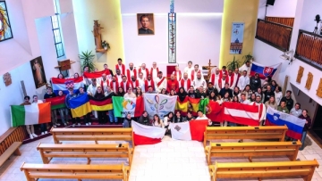 Circa 50 junge Menschen und Priester stehen in einer Kirche und halten internationale Fahnen in den Händen. Davor stehen drei Reihen Kirchenbänke.