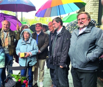 Sieben Männer und Frauen stehen mit Regenschirm auf einer Straße. Eine ältere Frau hält einen goldenen Stein in der Hand.