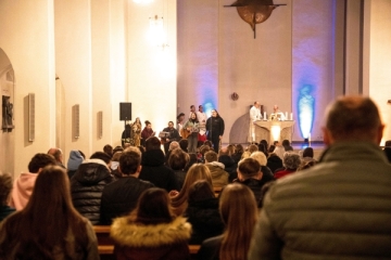In einer Kirche sind im Vordergrund Menschen in Kirchenbänken  von hinten zu sehen. Vorne steht eine Sängerin und am Altar zwei Priester in weißen Gewändern.