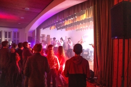 In einem dunklen Raum  stehen junge Menschen vor der B&uuml;hne. Auf der B&uuml;hne spielt eine Band, die von roten, violettenund blauen Scheinwerfern angeleuchtet wird.