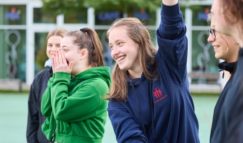 Vier Jugendliche stehen draußen vor einem Gebäude. Ihr Oberkörper . Ein Mädchen hebt ihren Arm hoch und lacht.