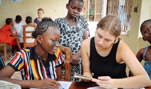 Eine Lernsituation: Ein Mädchen in Benin lernt und wird dabei von einer jungen Frau, die einen Freiwilligendienst als Don Bosco Volunteers macht, unterstützt. Um sie herum stehen zwei weitere Kinder, im Hintergrund sieht man eine andere Lerngruppe.
